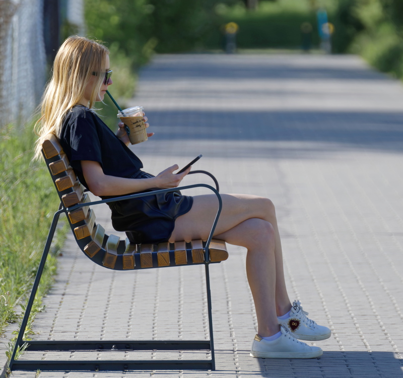 A blonde woman sitting on a modern outdoor bench while holding a drink and looking at her phone
